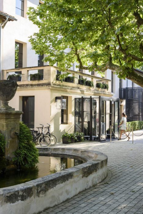 Audit hôtel : bâtiment élégant entouré de verdure, terrasse soignée, vélos stationnés, fontaine en pierre, ambiance calme et professionnelle, pénétrant la lumière naturelle à travers de grandes fenêtres.