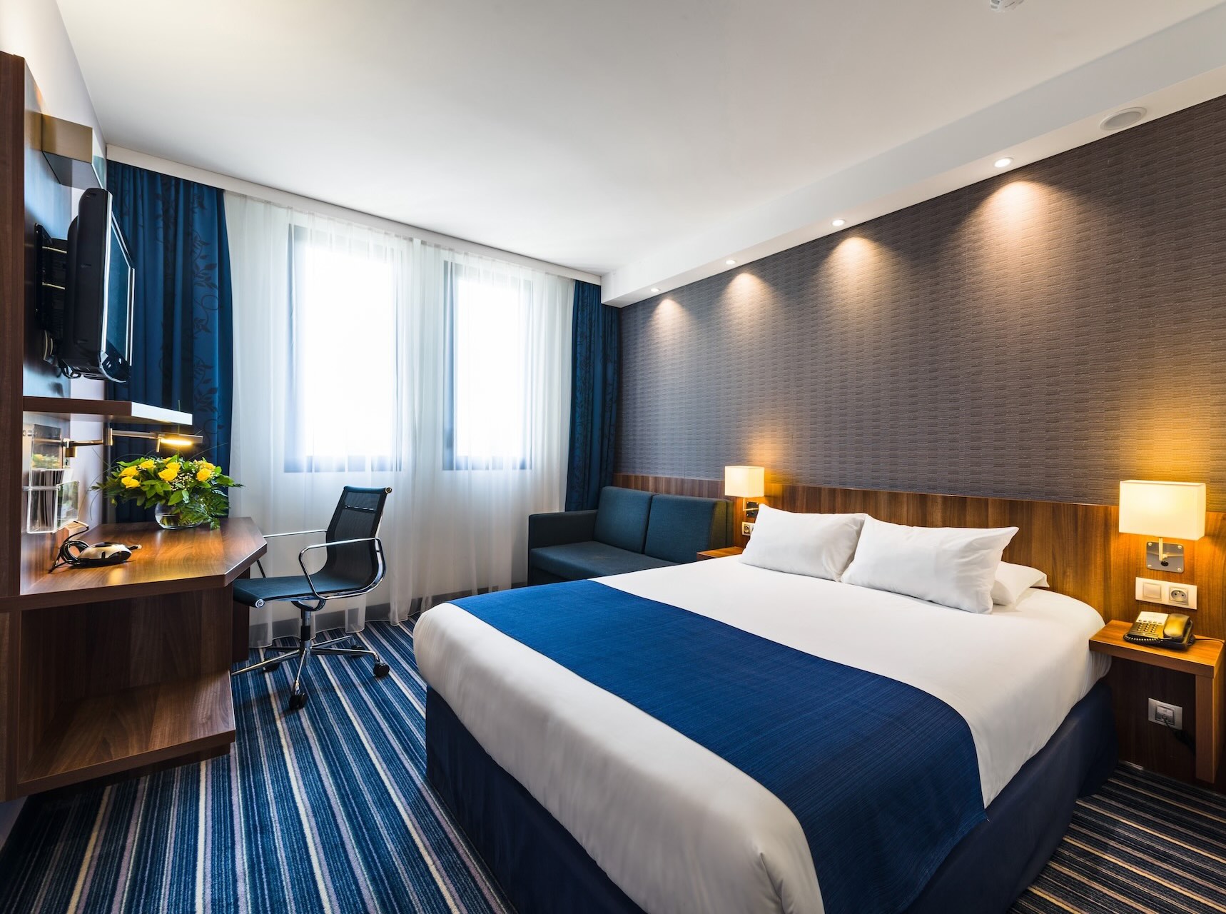 Chambre d'hôtel moderne avec lit double, draps blancs et couverture bleue, bureau avec chaise, canapé, deux lampes et tapis à rayures, éclairage naturel par deux fenêtres.